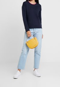 Pull à manches longues bleu marine avec un col rouge et blanc, jean droit bleu clair, baskets blanches et un petit sac à main jaune avec une poignée en métal.