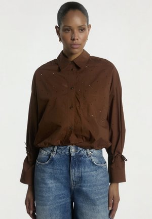 Vrouw draagt een bruin blouse met kleine stippen en blauwe high-waisted jeans, staand tegen een effen lichte achtergrond.