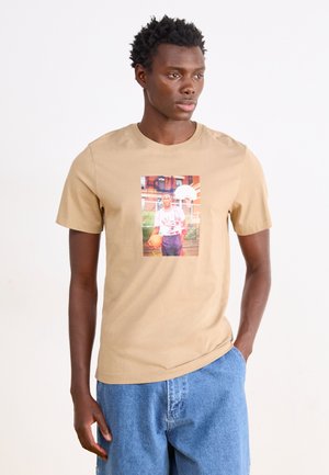 Tricou din bumbac bej cu un grafic central care prezintă o persoană ținând o minge de baschet, completat de pantaloni scurți din denim albastru.