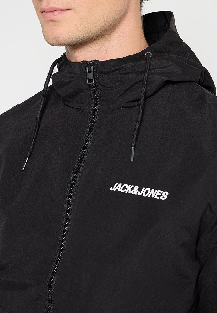 Veste zippée noire avec capuche, fabriquée en matériau léger. Elle dispose de cordons de serrage, d'un logo blanc sur la poitrine et d'une texture lisse.