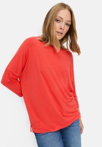 Rød langærmet top med afslappet pasform, draperet design og sideslidser, lavet af letvægtsstof. Kombineret med blå denim jeans.