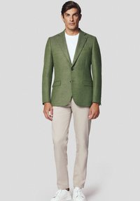 Zielona wełniana marynarka z dwoma przednimi kieszeniami, klapami, oraz z zamknięciem na jeden guzik, w zestawieniu z jasnobeżowymi spodniami i białymi sneakersami.