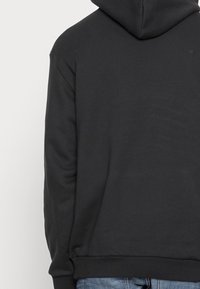 Vue arrière d'une personne portant un sweat à capuche noir uni et un jean bleu devant un fond neutre.