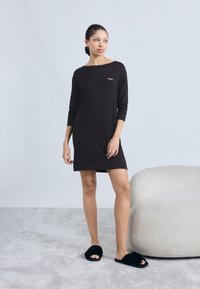 Black long-sleeve dress with a round neckline, featuring a small white logo. Model wears black fuzzy slippers, standing on a light gray carpet.