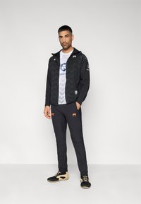 Veste noire zippée avec motifs géométriques, t-shirt graphique blanc, pantalon noir avec logo orange, et chaussures noires avec accents beige.