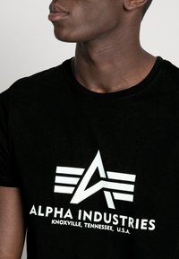 Muž v černém tričku s bílým logem Alpha Industries a textem "Knoxville, Tennessee, USA" na jednoduchém světlém pozadí.