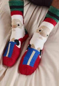 Sarkani un balti pūkaini zeķes ar Ziemassvētku vecīša dizainu, zaļiem un melniem svītrainiem galotnēm, un zilu un zeltu dāvanu detaļu uz pirkstiem.