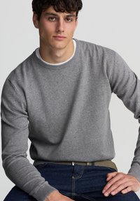 Maglione grigio lavorato a maglia con scollo rotondo e polsini a costine, abbinato a jeans blu scuro. Il tessuto appare morbido e testurizzato.