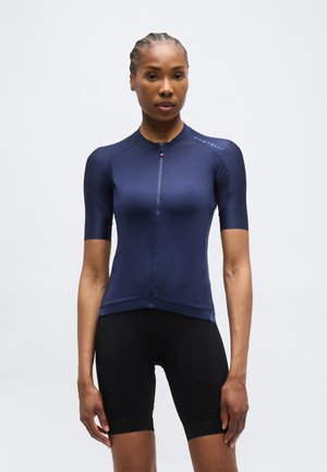 Γυναίκα με πλεξούδες, φορώντας μπλε ναυτικό ποδηλατικό μπλουζάκι με κοντά μανίκια και μαύρο ποδηλατικό σορτς, στέκεται μπροστά σε απλό φόντο.