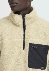 Fleece pullover in cream with a high collar, half-zip front, and a contrasting black chest pocket featuring a zipper and brand label.