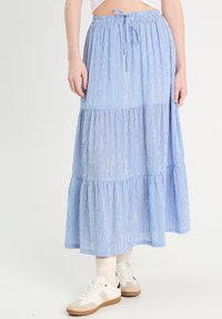 Jupe maxi rayée bleu clair avec un design à volants, dotée d'une taille à cordon de serrage et d'une texture de tissu douce. Portée avec des baskets blanches.