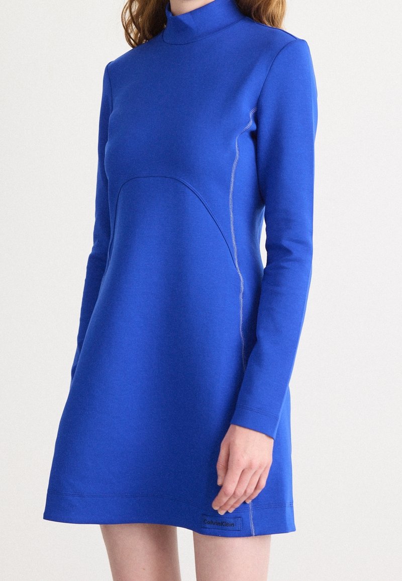 Femme portant une robe courte à manches longues, bleu roi, col montant, avec des détails subtils de coutures et un petit logo Calvin Klein près de l'ourlet.