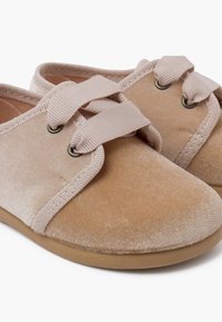 Scarpe slip-on in velluto beige con una parte superiore morbida e testurizzata, cinturini elastici e una suola in gomma flessibile. Presentano hardware a occhio per il fissaggio dei cinturini.