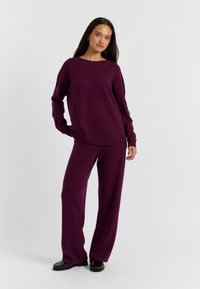 Ein pullover in Pflaumenfarbe und weit geschnittene Hose, mit langen Ärmeln und lässiger Passform. Das Material wirkt weich und hat sichtbare Nähte.