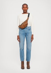 Leopard-print fanny pack with a smooth texture, black zipper, worn diagonally across a white sweater and blue high-waisted jeans. Brown ankle boots.