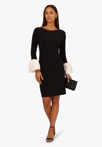 Czarna sukienka z długimi rękawami, z białymi, rozszerzającymi się mankietami; dopasowana sylwetka z marszczeniem z boku; zestawiona z czarną kopertówką i sandałami.