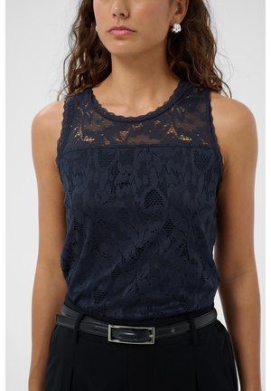 Femme portant un haut sans manches en dentelle bleu marine à motif floral, rentré dans un pantalon noir avec une ceinture en cuir noire.