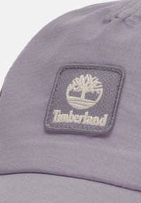 Jasnofioletowa czapka wykonana z teksturowanej tkaniny, z przeszywaną kwadratową łatą z logo drzewa i napisem "Timberland" na dole.