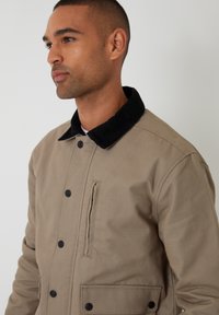 Beige jas met een zwarte corduroy kraag, drukknoopsluiten en een ritszak. Heeft een gladde textuur en een relaxte pasvorm.