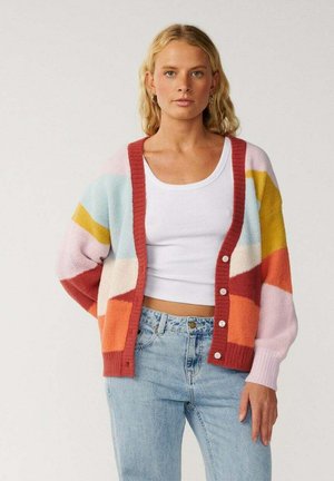 Jonge vrouw met golvend blond haar draagt lichtblauwe jeans, een wit cropped tanktop en een kleurrijk gestreept vest met knopen.
