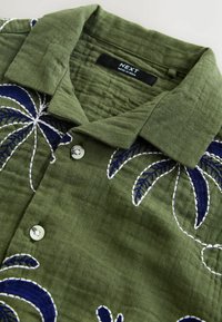 Olivgrünes Hemd mit Knöpfen, mit marineblauen und weißen bestickten Palmblattmuster und einem umgeschlagenen Kragen, Etikett mit der Aufschrift "NEXT Made in India."