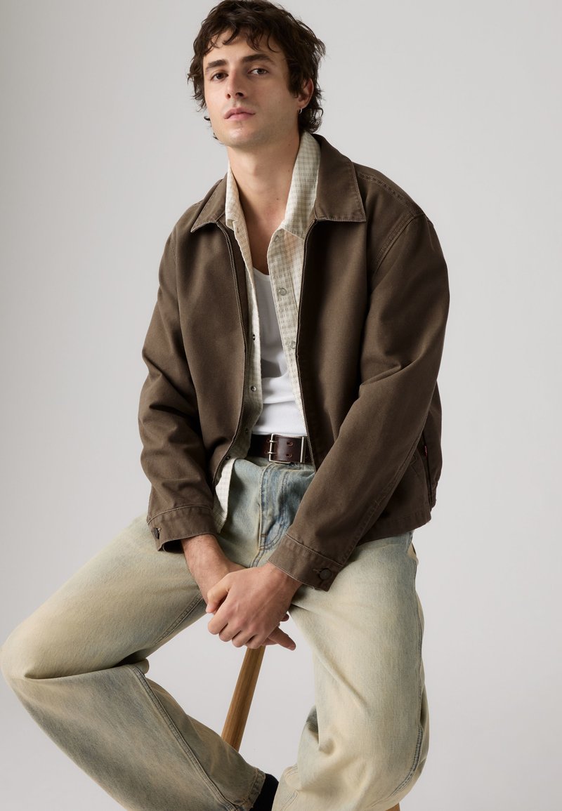 Joven sentado en un taburete con chaqueta marrón, camisa beige abotonada, camiseta blanca interior, vaqueros claros y cinturón oscuro sobre un fondo liso.