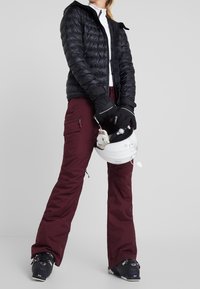 Svart puffjacka med huva, vinröda skidbyxor med dragkedjefickor, svarta handskar och en vit skid hjälm hålls i handen.