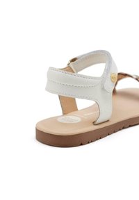 Sandalia de cuero blanco con suela plana marrón, que presenta dos correas y un botón decorativo dorado cerca del tacón. Textura suave, diseño minimalista.