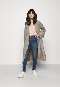 Casaco de xadrez longo em castanho e creme, blusa rosa com decote em V, jeans azuis de cintura alta e sapatos slip-on brancos com plataforma. Texturas suaves em toda a parte.