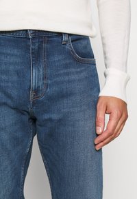 Close-up of person wearing blue denim jeans and a white long-sleeve top, with right hand relaxed by their side.