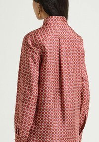 Blusa rossa a motivi con disegni geometrici, maniche lunghe e collo alto. Tessuto liscio con dettaglio di piega posteriore.