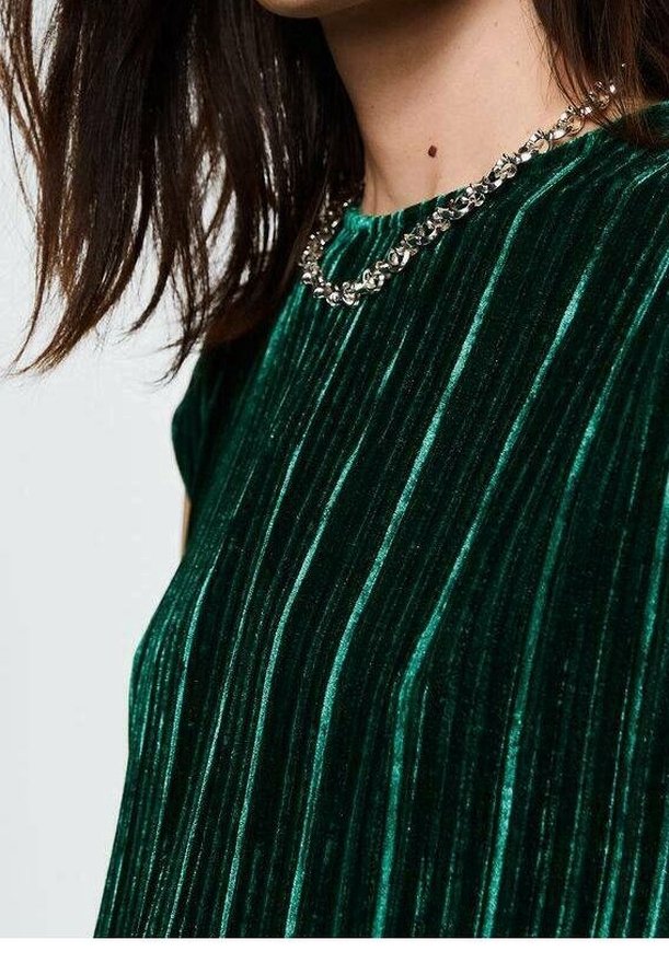 Femme portant un haut plissé en velours vert foncé et un collier chaîne argenté, avec des cheveux bruns mi-longs visibles.