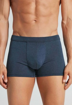 Torso masculino con bóxers ajustados de color azul oscuro, mostrando el abdomen y la parte superior de las piernas contra un fondo claro y liso.