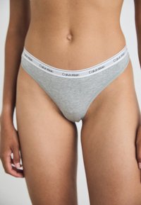 Sivi pamučni tanga gaćice s bijelim pojasom na kojem je logo "Calvin Klein". Glatka tekstura i niska visina.