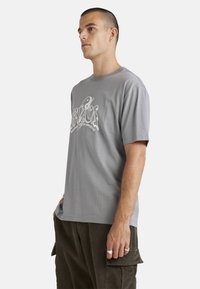 Gray cotton t-shirt with short sleeves, featuring a graphic design on the front. Model wears corduroy pants, showing a relaxed fit.