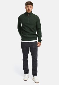 Mann som står iført en mørk grön glidelåsgenser, hvit t-skjorte, svarte jeans og hvite sneakers mot en ensfarget bakgrunn.