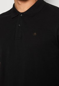 Polo noir en tissu texturé avec une patte de boutonnage à trois boutons et un petit logo doré sur la poitrine.