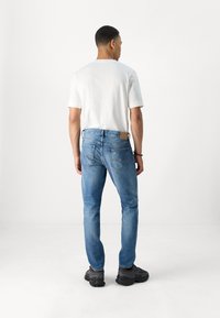 Vaqueros de mezclilla azul con corte slim, con un parche de cuero marrón en la parte trasera de la cinturilla, combinados con una camiseta blanca y zapatillas negras.