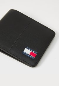 Black leather wallet with stitched edges, featuring a rectangular Tommy Jeans logo in blue, white, and red on the front.