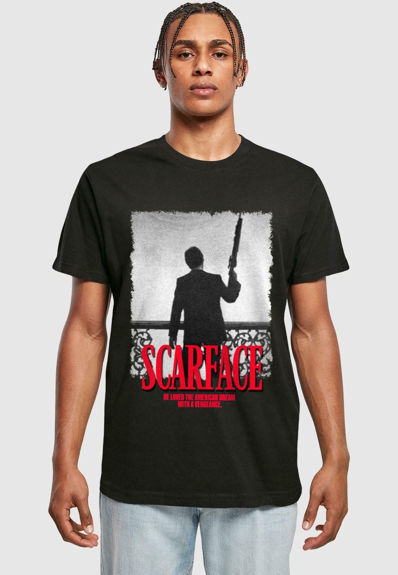 Man wearing black t-shirt featuring black-and-white silhouette holding rifle and red "Scarface" text with tagline underneath.