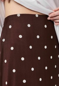 Primo piano di una persona che solleva una camicia bianca, rivelando una gonna marrone con un motivo a pois bianchi intorno alla vita.