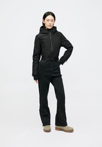 Fato de esqui isolante preto com um design ajustado, capuz, cintura cingida e bolsos com fecho. Combinado com botas de inverno bege.