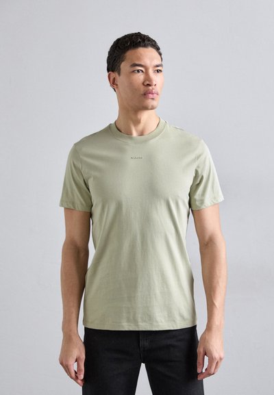 T-shirt en coton vert clair avec un col rond, des manches courtes et un logo de marque subtil centré sur la poitrine. Porté avec un pantalon noir.