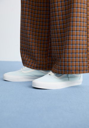 Des baskets bleu clair portées avec un pantalon à carreaux marron et bleu à jambes larges sur un sol en tapis bleu clair.