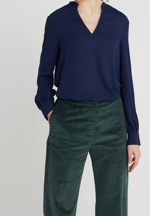 Personne portant un chemisier bleu marine à manches longues avec un col en V, rentré dans un pantalon en velours côtelé vert foncé à taille haute, une main dans la poche.
