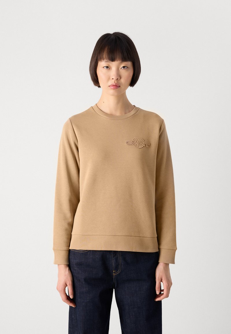 Frau in einem hellbraunen langärmeligen Sweatshirt mit einem kleinen Brustlogo, kombiniert mit dunkelblauen Jeans, steht vor einem schlichten hellen Hintergrund.