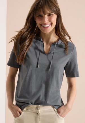 Uśmiechnięta kobieta o długich brązowych włosach, ubrana w szarą bluzę z krótkim rękawem i beżowe spodnie, z rękami w kieszeniach, na jednolitym tle.