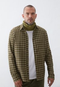 Grønternet langærmet skjorte med sorte knapper, lagd over en hvid T-shirt med rund hals. Olivengrøn halsvarmer. Struktureret stof, løs pasform.
