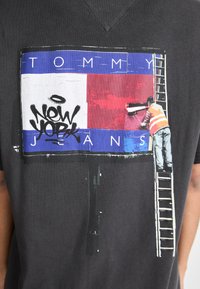 Camiseta negra de algodón con un gráfico de un pintor en una escalera, pintando una valla publicitaria con secciones en rojo y azul, etiquetada "TOMMY JEANS."
