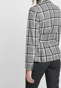 Blazer à carreaux gris avec des lignes noires et blanches, coupe ajustée, tissu texturé et taille définie. Coupe slim, associé à un pantalon à rayures noires.